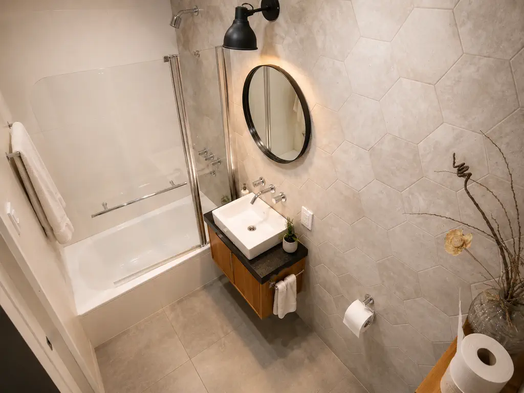 Baño con bañera y mampara de vidrio, revestimiento hexagonal, espejo redondo y bacha de apoyo