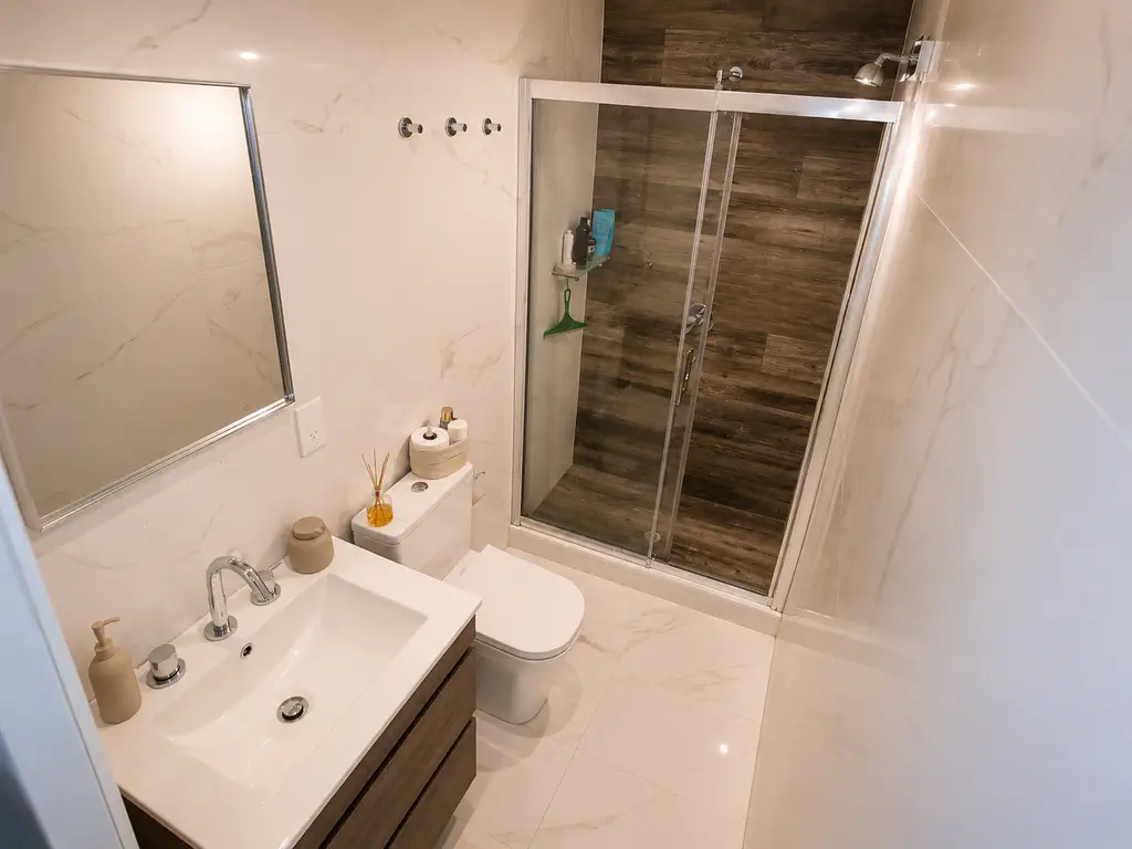 Baño en suite con ducha y mampara de vidrio corrediza, revestimiento símil mármol y vanitory de madera